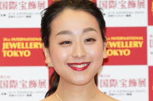 女性が、シンプルなネックレスと肩の露出がある黒いドレスを着て、笑顔でポーズを取っています。バッグには赤いロゴ。