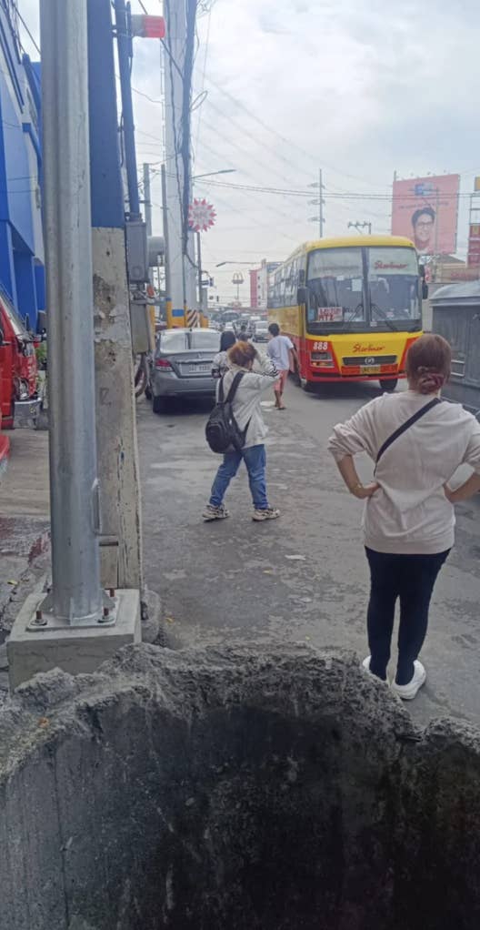 Two people stand on a busy street near a large pothole, with traffic and a bus in the background