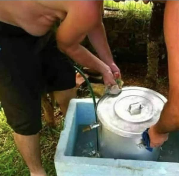 Two people outdoors washing a large metal pot with a hose and sponge