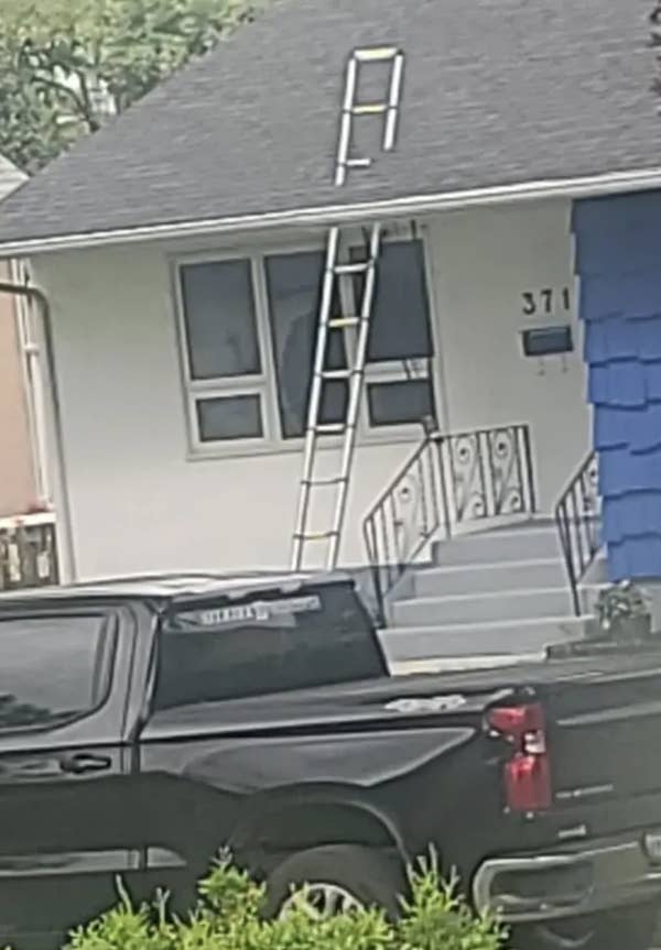 A ladder is precariously positioned on both a truck bed and roof, reaching from the ground to a house roof