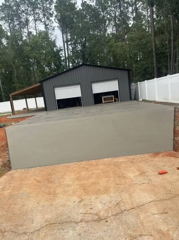 A new concrete driveway leads to a gray garage with two doors, surrounded by trees and a white fence