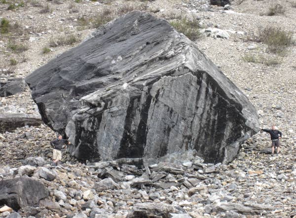 Two people push against a large, jagged boulder on a rocky landscape, showcasing the enormity of the rock