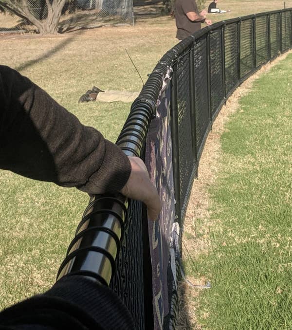 A person leans on a curved fence in a sunny outdoor park, grass visible, another person on the ground in the background