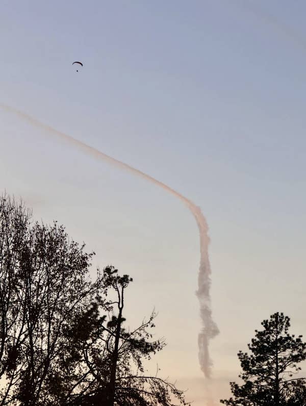 Sky with contrail and drifting smoke, silhouettes of trees in foreground; a distant parachute visible above