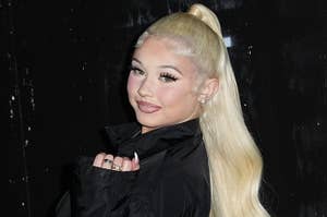Alabama Barker with long hair in a high ponytail, wearing a stylish black coat, poses smiling against a dark background