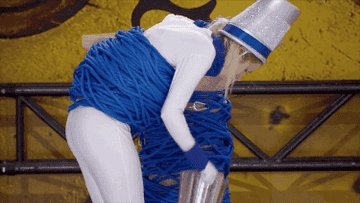 Person in a comedic costume wrapped in blue ropes, wearing a metallic bucket hat, leaning over