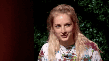 Person with long hair smiles and talks, wearing a tie-dye shirt. Background is greenery