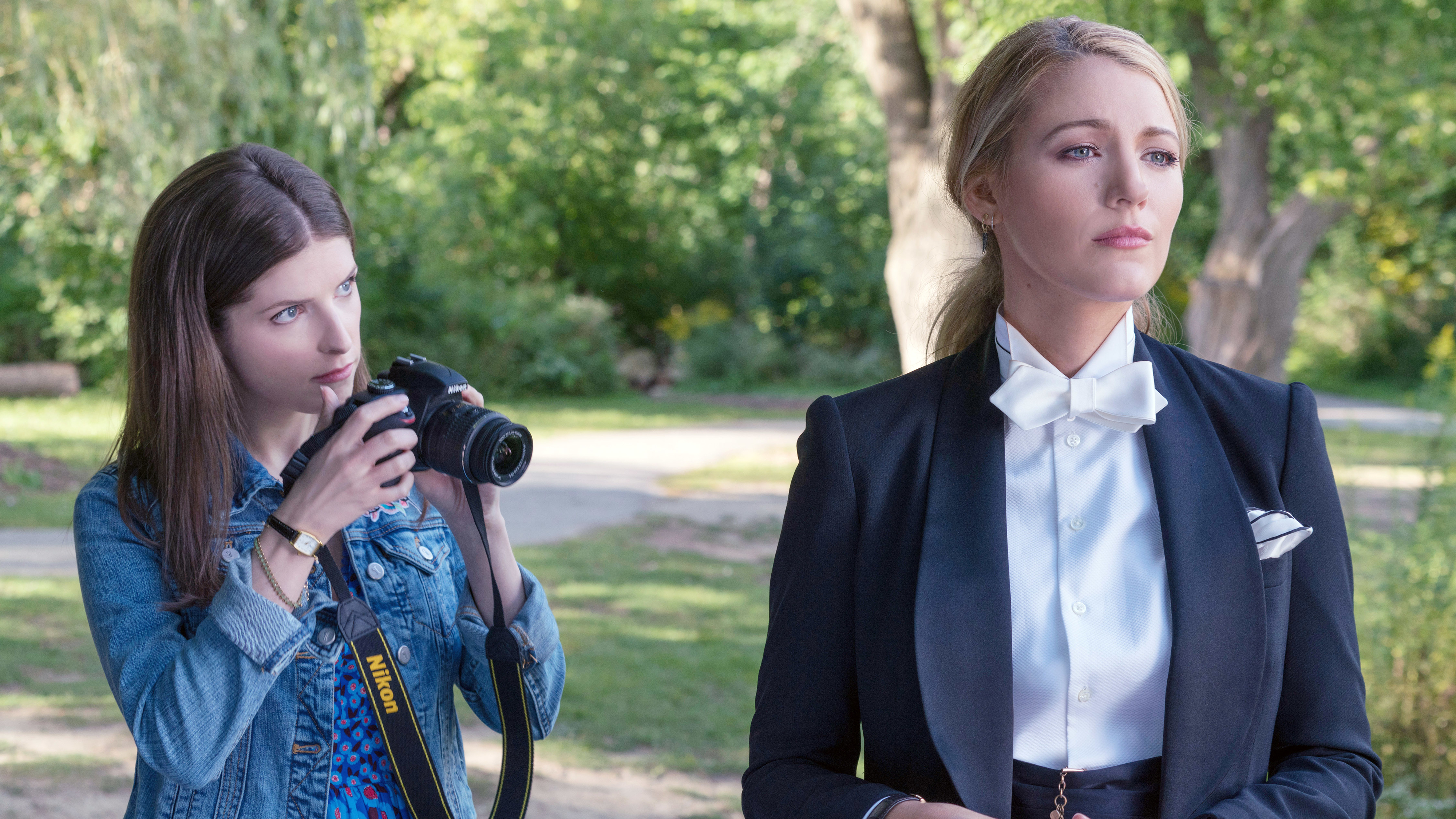 Two individuals outdoors: one with a camera, the other in a formal suit with a bow tie. From a scene in a TV or movie setting