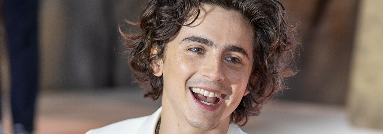 Timothée Chalamet smiling in a white suit with wavy hair, standing on a carpeted area at a formal event
