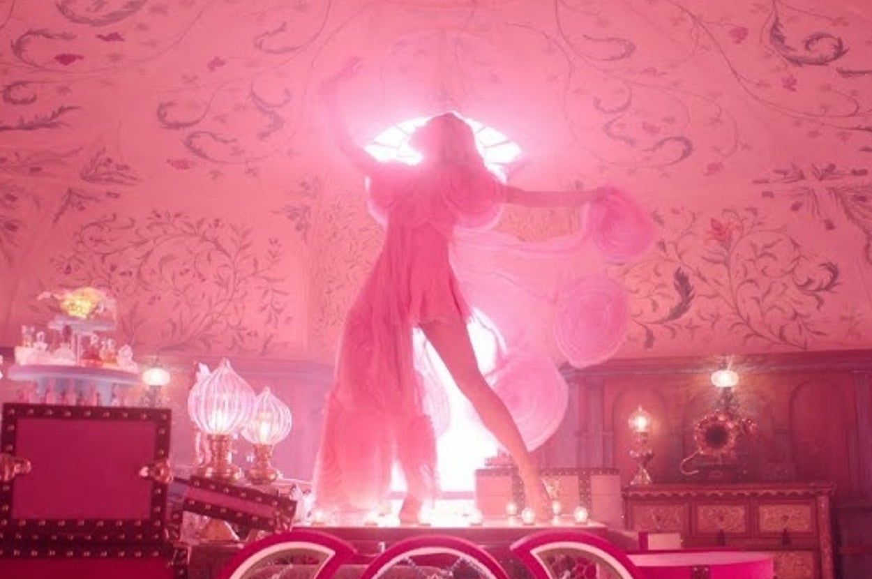 Person in flowing sheer dress poses dramatically on a platform in an ornate room with intricate wall designs and soft lighting