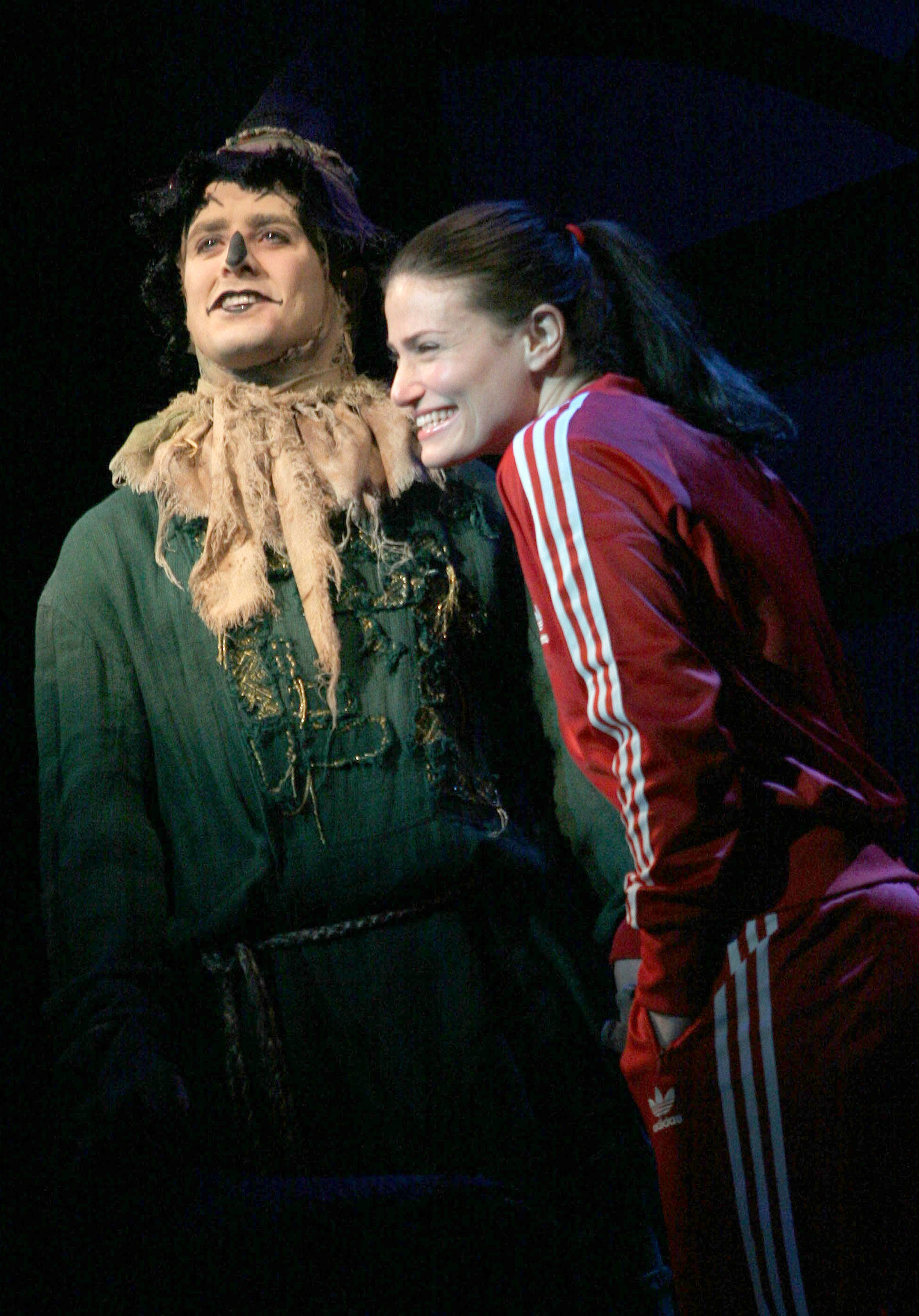 Idina Menzel smiling wearing a red tracksuit while next to an actor playing Fiyero in Wicked