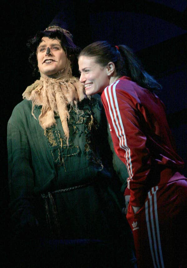 Idina Menzel smiling wearing a red tracksuit while next to an actor playing Fiyero in Wicked