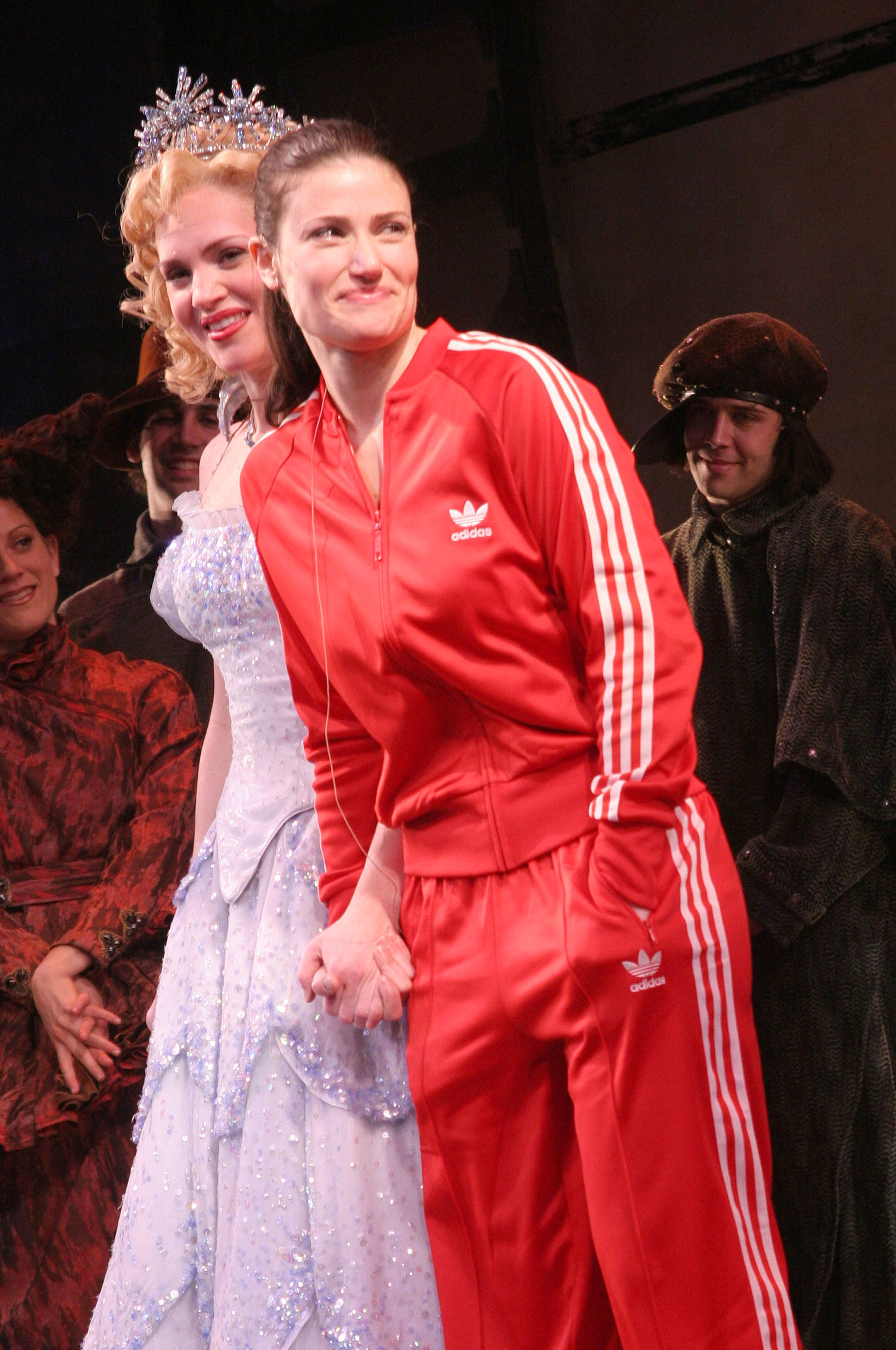 Idina Menzel in a red tracksuit holding hands with an actor playing Glinda on stage at Wicked