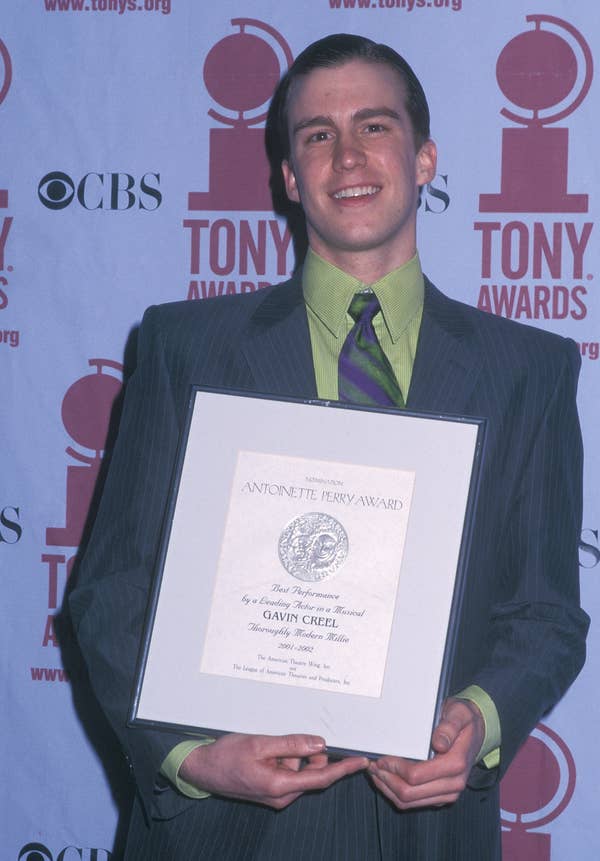 Gavin Creel smiling in pinstripe suit, holding a framed certificate at a Tony Awards event backdrop in 2002
