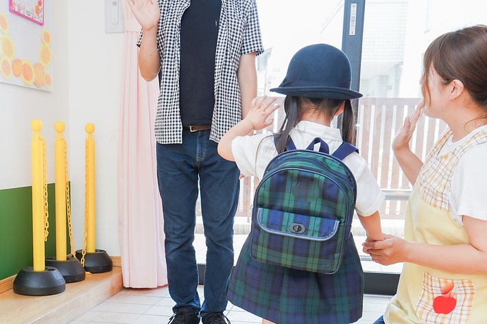 幼稚園で起立する女の子が保護者と先生に手を振っている場面。女の子は制服とリュックを身に着けている。