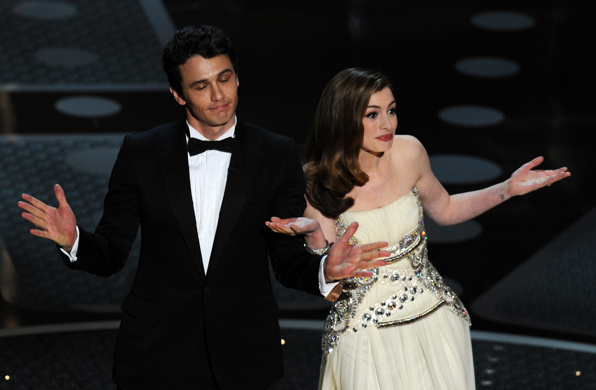 Two people on stage shrugging. James Franco wears a tuxedo; Anne Hathaway is in an embellished white gown