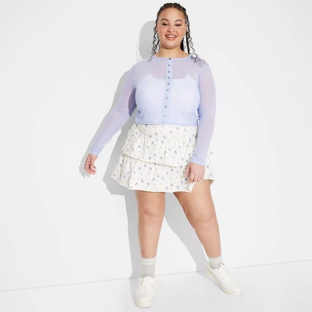 Person in a stylish, sheer-sleeved blouse and floral tiered skirt, standing confidently in casual white sneakers