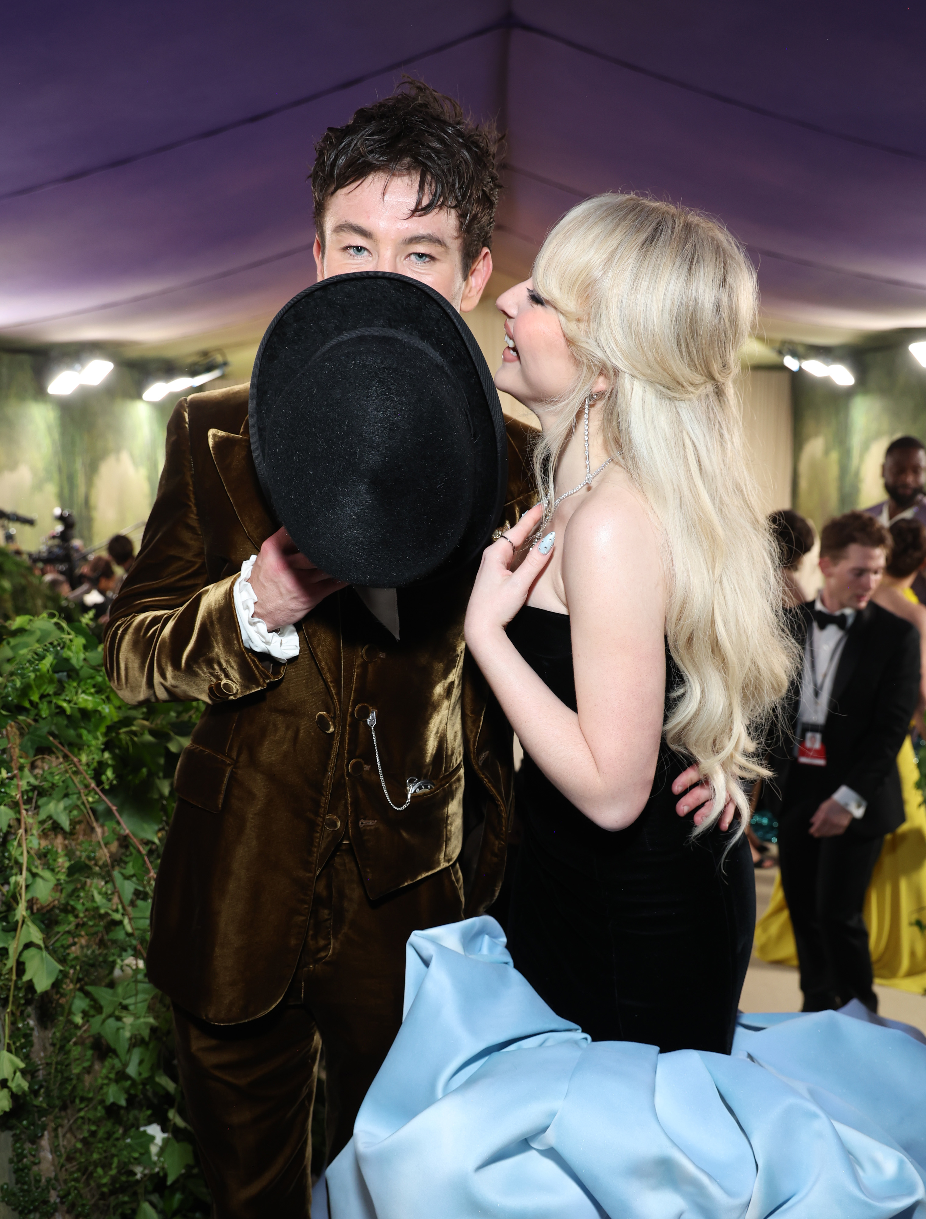 Two people on a red carpet; one in a velvet suit holding a hat and the other in a strapless gown, sharing a candid, joyful moment