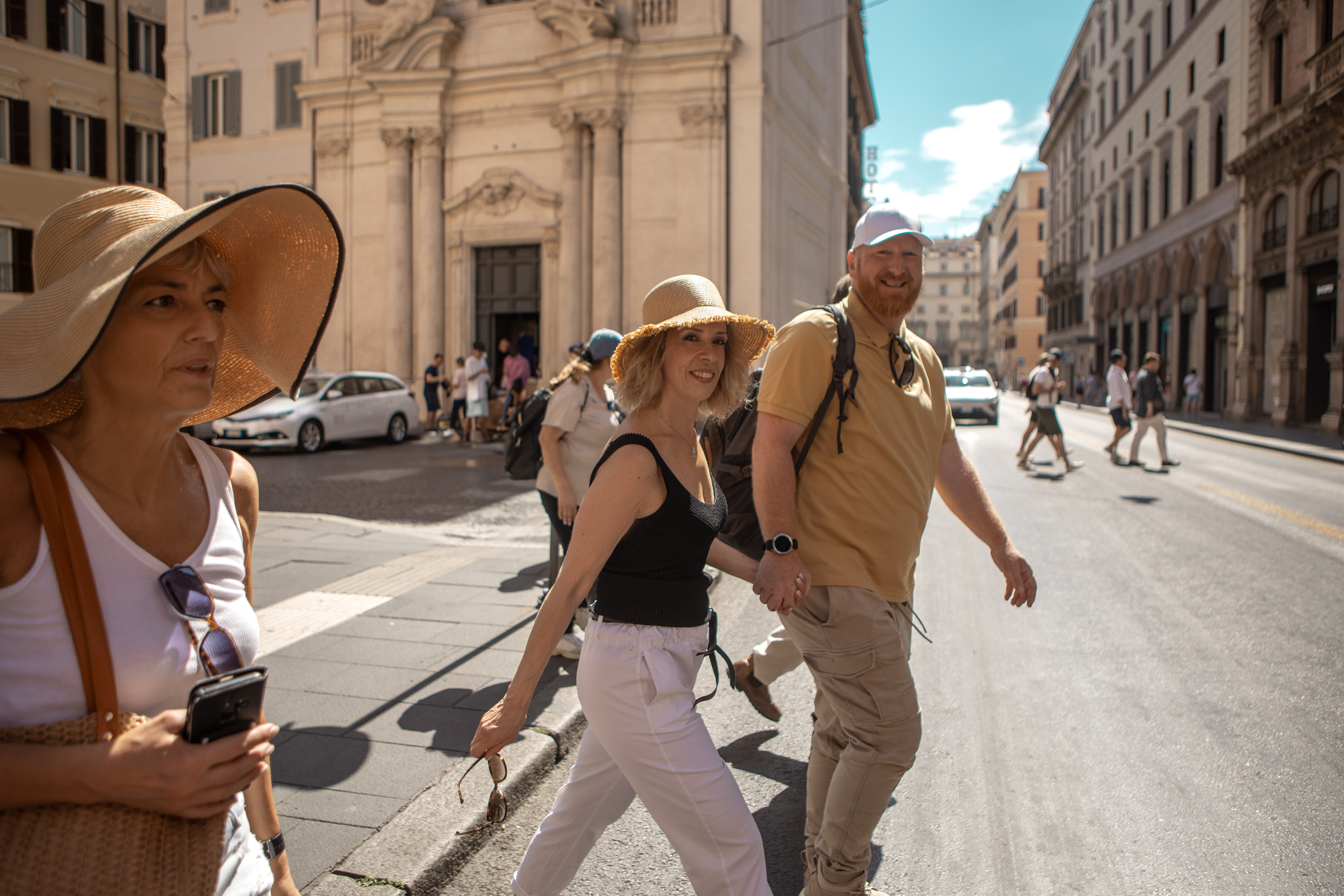 People crossing a street, wearing casual summer clothing and hats, in a historic city setting