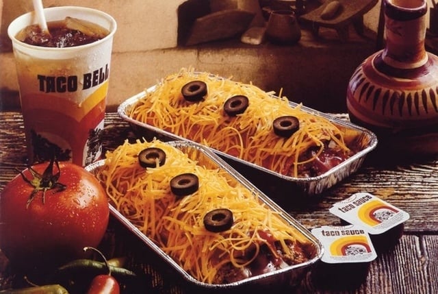 Vintage Taco Bell meal with 2 Taco Bell grande meals topped with shredded food and olives, a drink, and taco condiment packets connected a woody table