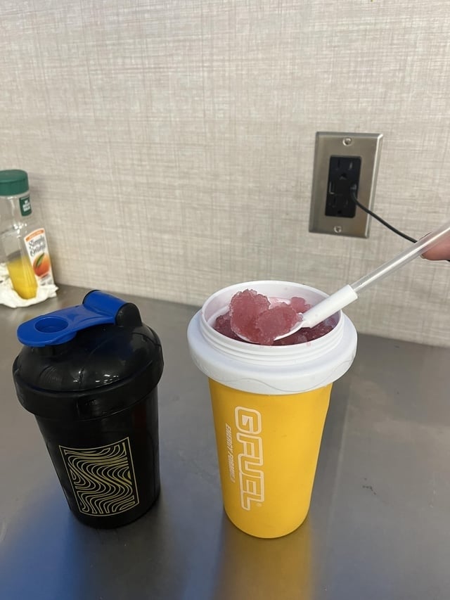 A yellowish G FUEL shaker cupful with reddish slushy inside, adjacent to a closed achromatic shaker vessel connected a counter