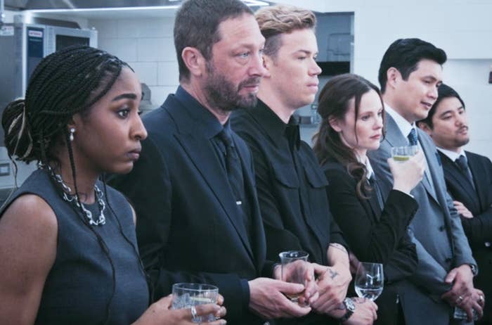 Group of six people in formal attire standing together, holding drinks, with serious expressions. They appear to be in a professional setting