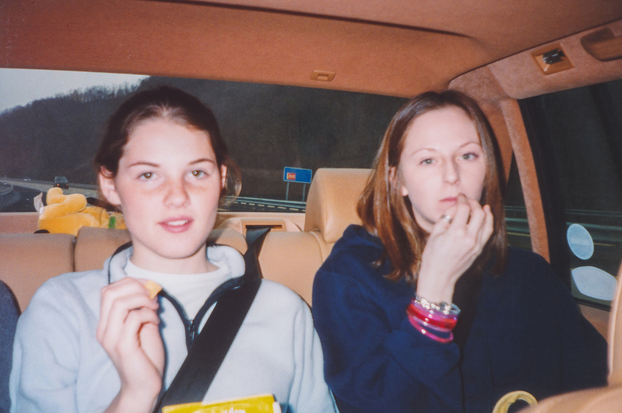 Two people sitting in a car, eating snacks and looking at the camera. A plush toy is in the background on the seat