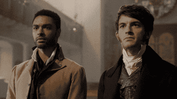 Two men dressed in period costumes, possibly from a historical drama, stand together looking ahead in an ornate interior setting