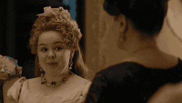 Woman in period costume with elaborate hairstyle and jewelry, engaged in conversation at a formal event