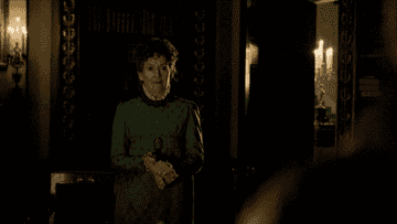 Elderly woman in an ornate green dress with white lace collar standing in a dimly lit, vintage room, holding a handkerchief