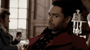 Man in stylish formal attire, sitting confidently in an elegant room, glancing sideways. Another person holds a drink in the background