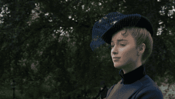 Person in a stylish outfit with a distinctive hat, outdoors in a leafy area, raising an eyebrow