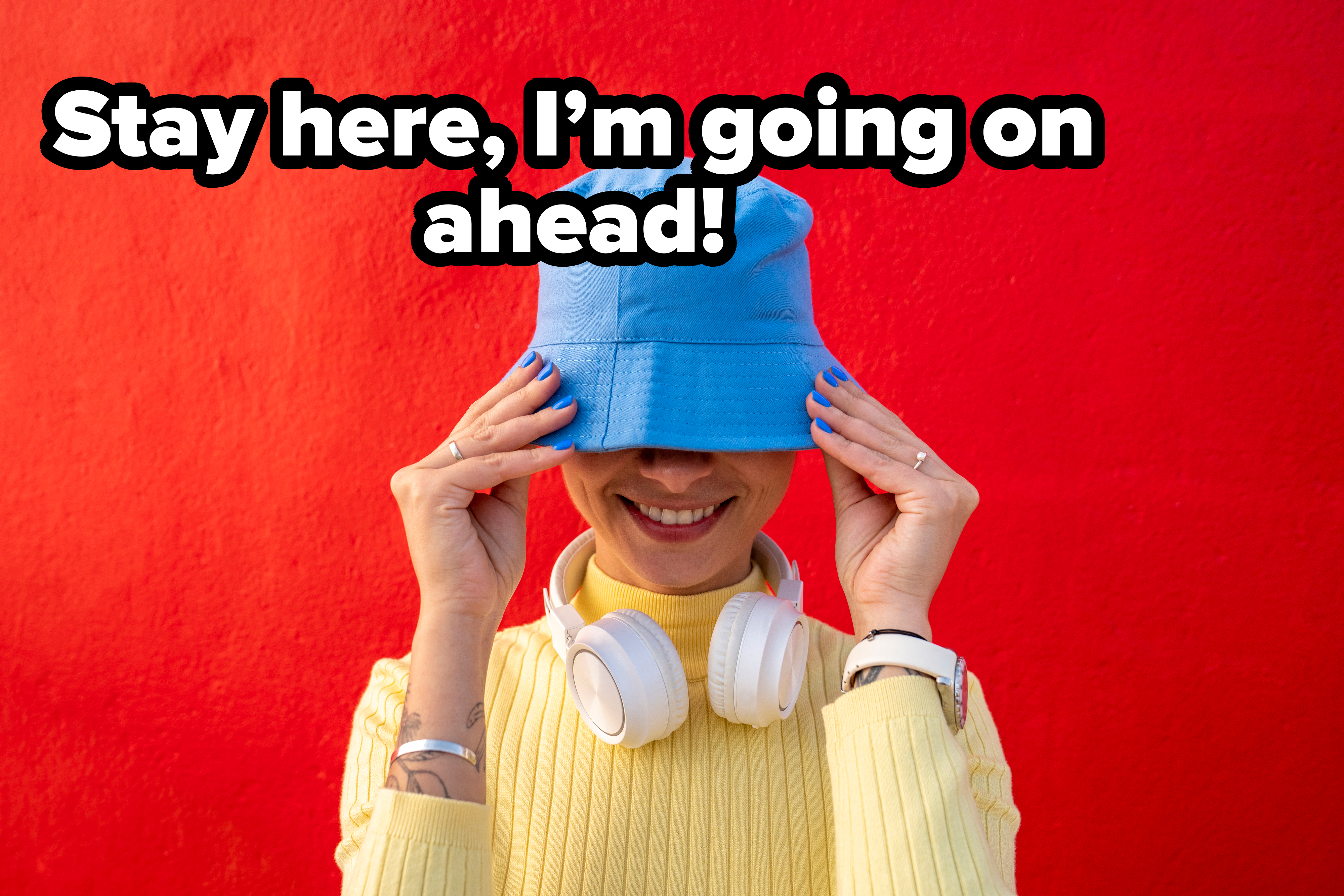 Person smiling, wearing a blue bucket hat and yellow sweater, with headphones resting around their neck against a vibrant red backdrop