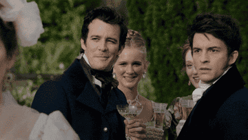 Group of people dressed in 19th-century formal attire holding champagne glasses, smiling and chatting at an outdoor event