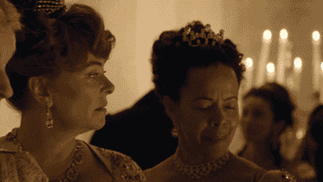 Two women in elegant, historic-style dresses and tiaras are engaged in conversation at an ornate, candlelit event