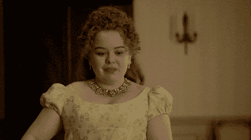 Person with curly hair and elegant necklace in a vintage-inspired gown, smiling and holding a sword between her hands