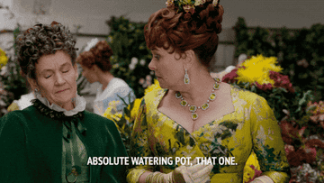 Two people in elaborate historical costumes converse at a floral event. Subtitles read: &quot;Absolute watering pot, that one.&quot;