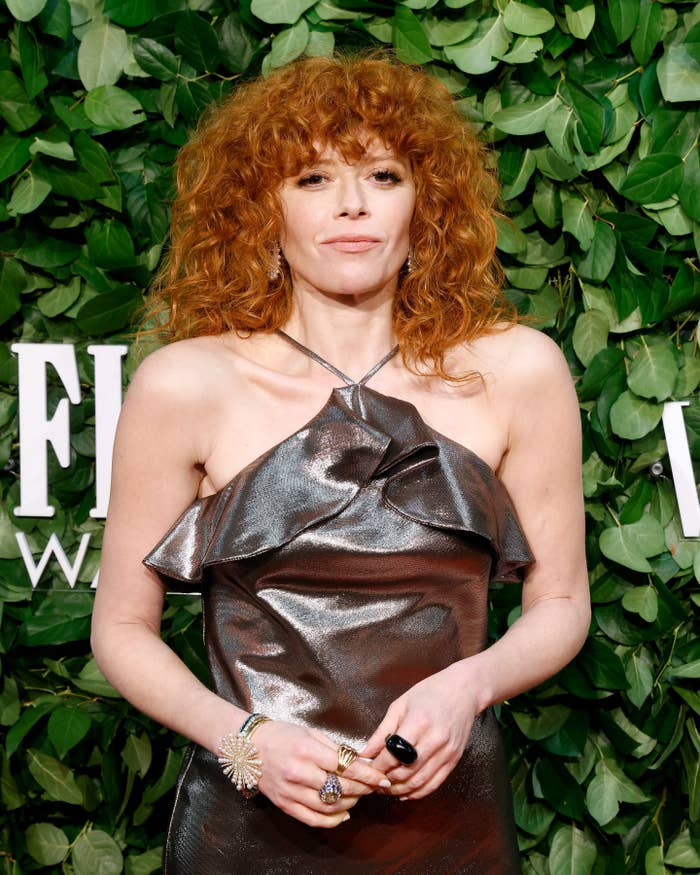Person with curly hair wearing a shiny, ruffled off-shoulder dress, posing on a leafy backdrop