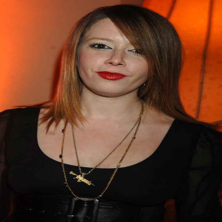 Natasha Lyonne, with straight, shoulder-length hair wearing a dark dress and layered necklaces, standing in front of soft, warm lighting