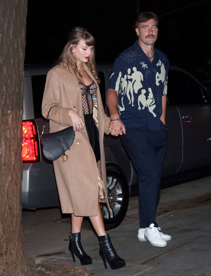 Taylor Swift in a stylish coat, lace top, and boots holds hands with Travis Kelce in a printed shirt and sneakers on a night out