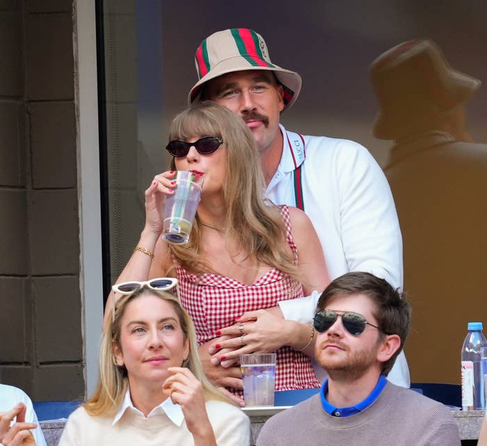 Taylor Swift wears a gingham dress, while Travis Kelce is in a jacket and bucket hat, holding her from behind