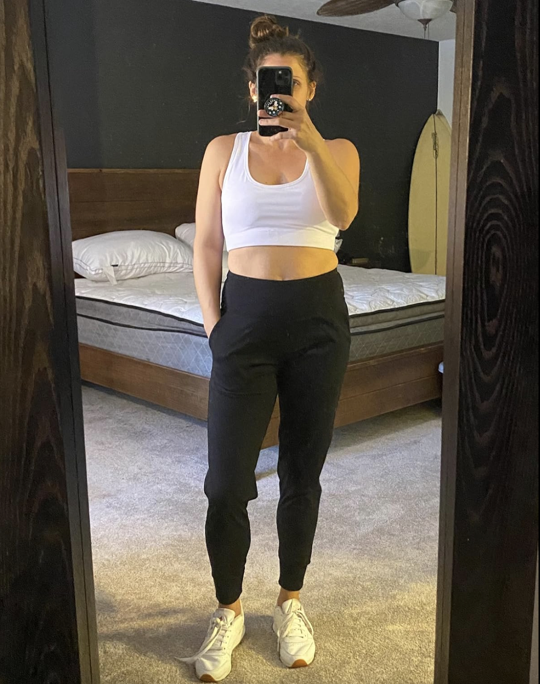 Person taking a mirror selfie in a bedroom, wearing a white tank top, black pants, and sneakers