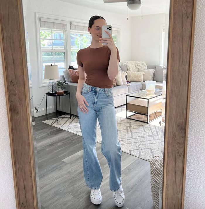 Person taking a mirror selfie in a casual living room, wearing a fitted top and wide-leg jeans with sneakers