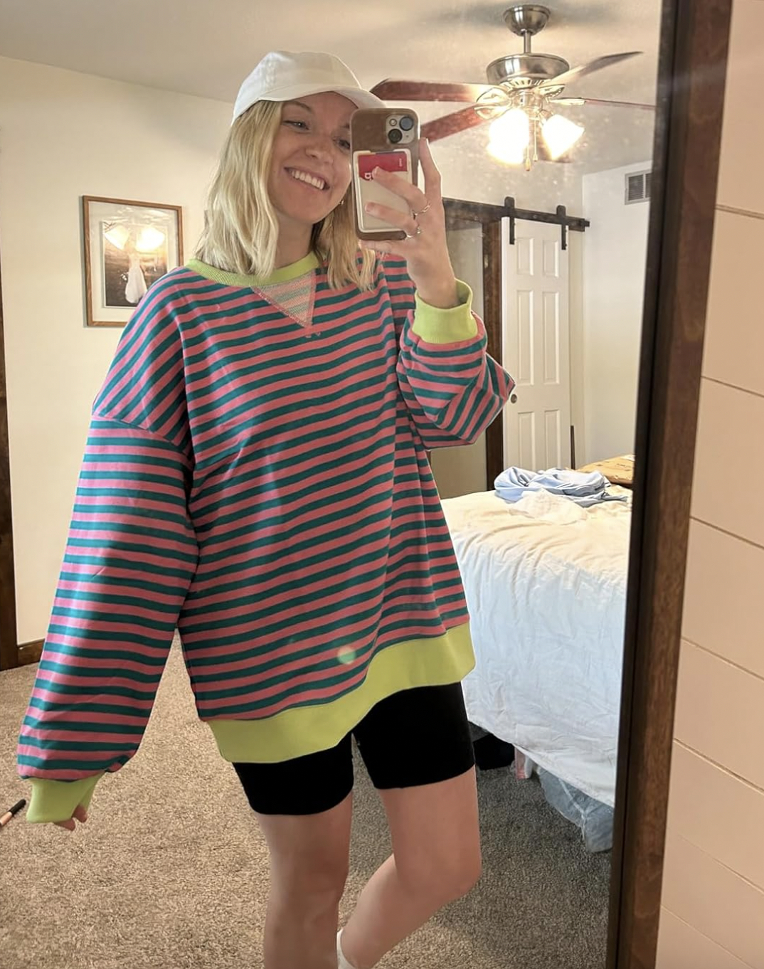 Person in striped oversized sweater and shorts takes a mirror selfie, smiling and wearing a cap. Bedroom setting with bed and ceiling fan visible