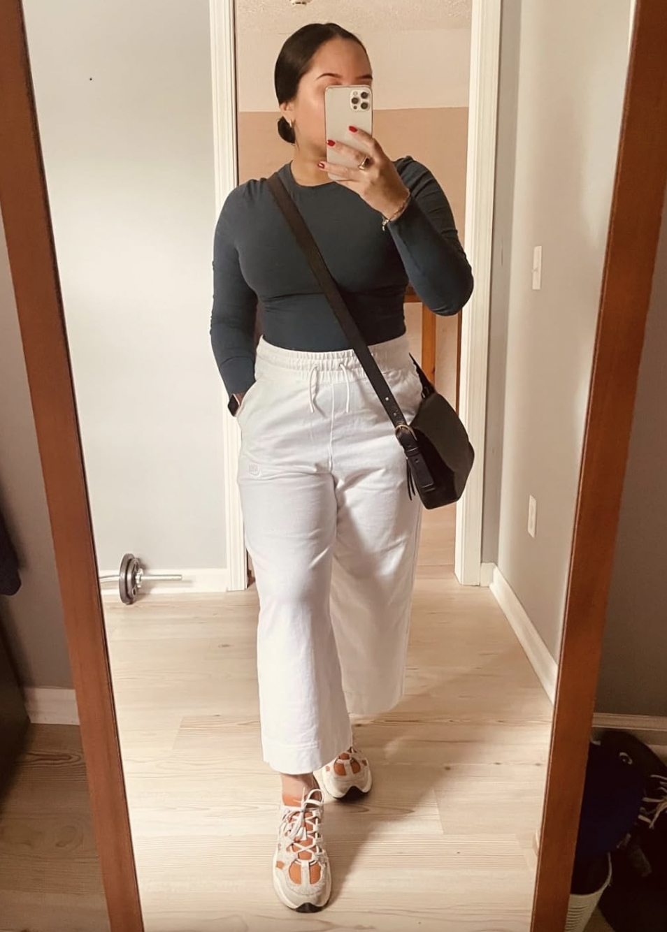 Person taking a mirror selfie, wearing a dark long-sleeve top, white wide-leg pants, sneakers, and a crossbody bag