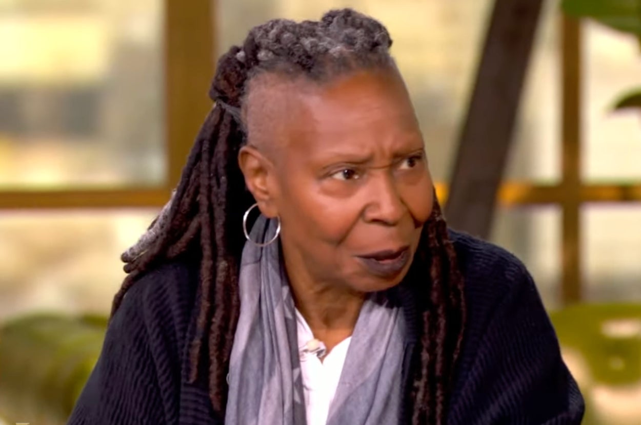 Person with long dreadlocks, wearing hoop earrings, a dark sweater, and a light scarf is speaking indoors