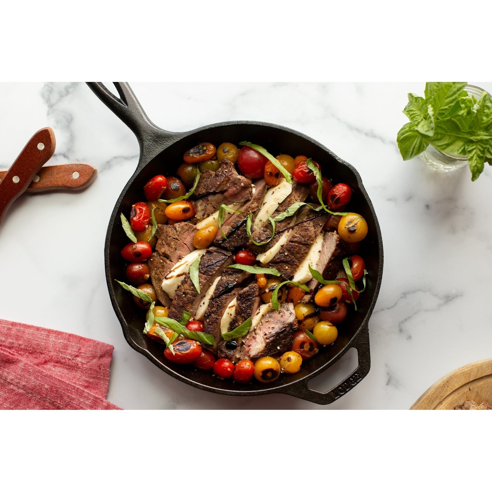 Skillet with grilled steak, cherry tomatoes, and herbs connected  a marble surface. Knife, napkin, and basil nearby