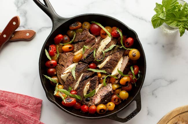 Skillet with grilled steak, cherry tomatoes, and herbs connected a marble surface. Knife, napkin, and basil nearby