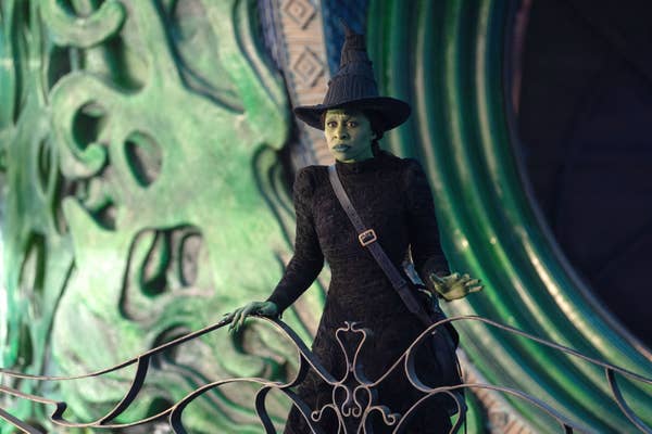 Person dressed as a witch with a pointed hat and black outfit, standing on an ornate balcony in a theatrical setting