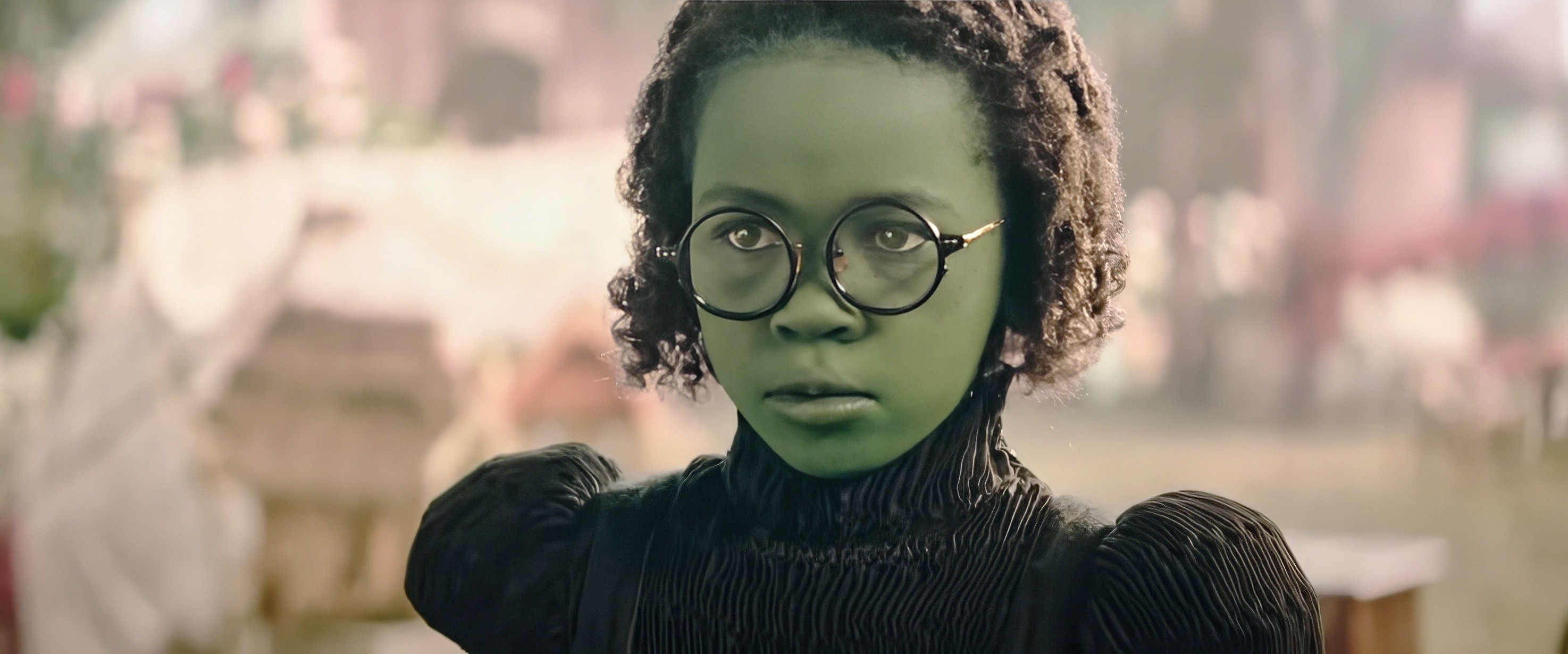 A person with green skin and round glasses wears a vintage-style black dress, appearing in a dramatic or fantasy setting